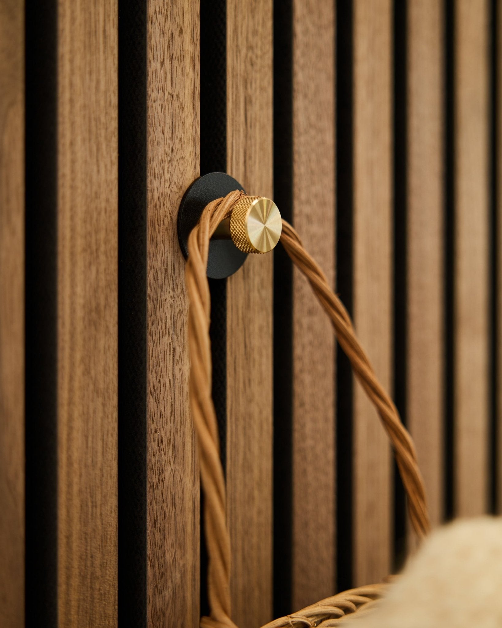 WoodUpp Norr Kleiderhaken in black und brass, montiert auf einer hölzernen Akupanel Akustikpaneel, hält einen geflochtenen Griff in Hellbraun.
